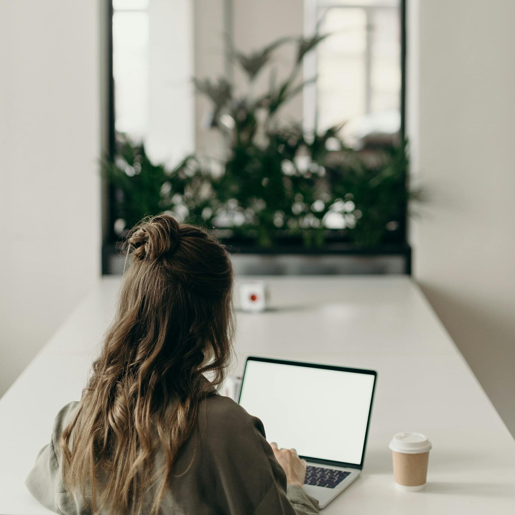 A woman with curly hair works on a laptop, facing a window with plants, symbolizing remote work.