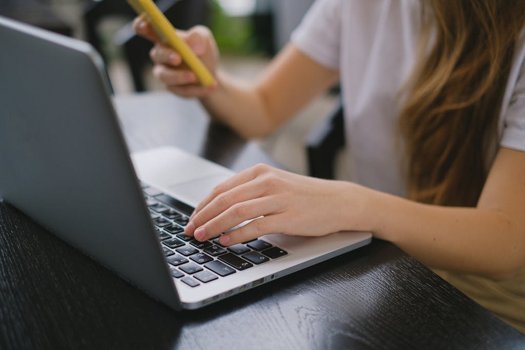 A young woman multitasks by using a laptop and smartphone indoors, focusing on work.