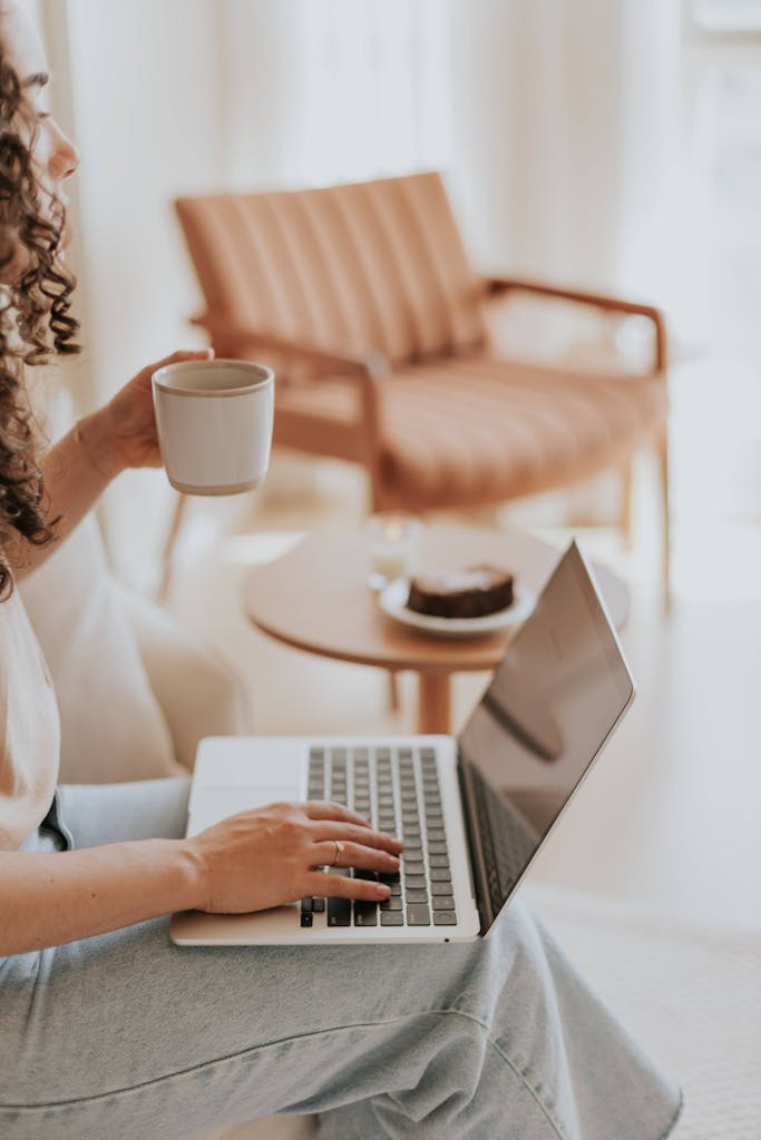 Woman working on a laptop in a cozy living room setting, sipping coffee, and enjoying a relaxing afternoon.